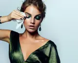 Mujer con maquillaje dramático verde, quitándose el rímel a prueba de agua con una toallita.