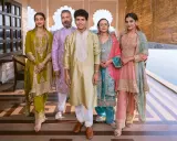 A family poses in vibrant traditional clothing from India. The women wear ornate salwar kameez sets in olive green, teal, and pink, while the men are dressed in elegant kurtas.