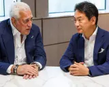 Koji Watanabe conversa con un hombre mayor, ambos con trajes azules. Un modelo de coche deportivo negro se ve en el fondo.