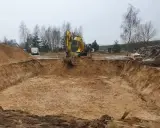 Koparka pracuje przy wykopy pod fundamenty. Widać głęboki dół w ziemi, usypane hałdy piasku i drzewa w tle.