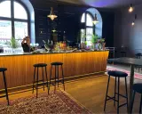 En bar med träpanel och svarta barstolar. En person bakom baren förbereder en drink. En meny på svarta tavlan visar priser för drycker i det musikaliska kvarteret.