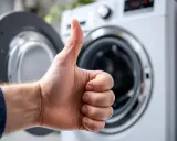 Pouce levé devant une machine à laver ouverte, signe que l'arrivée d'eau est parfaite pour le prochain lavage.