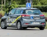 Szara Skoda i20 z oznaczeniami "Szkoła Jazdy EXPERT" i literą "L" na dachu i bokach. To auto do nauki jazdy, idealne dla przyszłego kierowcy.