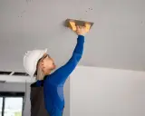 Kobieta w kasku i okularach wykonuje tynkowanie sufitu przy użyciu kielni.