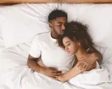 Ein Paar schläft eng zusammen in einem Bett. Er hält sie schützend im Arm, während sie sich an ihn kuschelt.