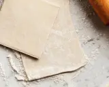 Pâte feuilletée prête à être travaillée, saupoudrée de farine sur un plan de travail en marbre. Un rouleau à pâtisserie en bois est visible sur la droite.
