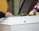 Dłoń z obrączką kładzie róże na białej trumnie, ozdobionej bukietem kwiatów i krzyżem.