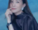 A woman with a thoughtful expression, her finger near her lips, wears a dark satin dress, a striking earring, and a blue-faced watch. She looks xa thi man.