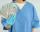Higienistka stomatologiczna w niebieskim fartuchu pokazuje wachlarz banknotów 20 dolarowych. Zarobki netto mogą być satysfakcjonujące.