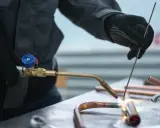 Ręce w rękawiczkach wykonują lutowanie gazowe miedzianych rurek. Płomień palnika rozgrzewa metal.