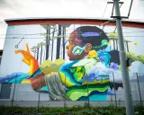 Färgstark street art på en vägg visar en person i profil med en fjäder i håret, omgiven av träd och blommor.