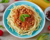 Pyszne spaghetti z bogatym sosem bolognese, udekorowane listkiem bazylii. Obok czosnek, pomidorki i przyprawy.
