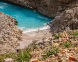 Malownicza zatoka z turkusową wodą i piaszczystą plażą, otoczona skalistymi klifami. Dwie osoby spacerują po brzegu. Chania plaże zachwycają.