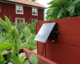 En solcellsdriven bevattning är monterad på en röd träkista i en trädgård, med ett rött hus i bakgrunden.