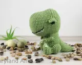A green crocheted dinosaur sits on a white surface surrounded by small stones and a small plant.