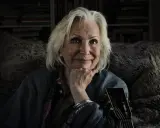 Äldre kvinna med grått hår, gitarr och en fluffig pläd. Trots att Marie Bergman är sjuk, lyser hennes ögon av liv.