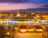 Der belebte marrakesch marktplatz erwacht bei Dämmerung zum Leben. Stände mit bunten Waren, Essensverkäufer und Menschenmassen beleben die Szene unter dem lila Himmel.