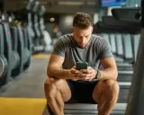 Mężczyzna w siłowni patrzy na telefon, siedząc na bieżni.