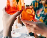 Zwei Hände halten Aperol Spritz Gläser mit Eis und Orangenscheiben. Ein perfektes Aperol Rezept für den Sommer.