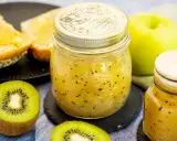 Ein Glas mit leuchtend gelber kiwi-marmelade mit schale, daneben frische Kiwischeiben und geröstetes Brot.
