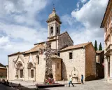 Piazza a San Quirico con una scultura moderna a forma di croce riflettente davanti alla chiesa romanica.