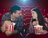 Para w kinie, zajadając popcorn, patrzy na siebie z uśmiechem. Ich spojrzenia sugerują, że to nie tylko film ich interesuje, ale też wzajemna bliskość.