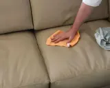 Mano che pulisce un divano in ecopelle beige con un panno arancione. Un altro panno grigio è appoggiato sul divano.