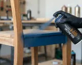 Main d'une personne portant un gant noir, peignant du mélaminé bleu sur un siège de chaise en bois avec une bombe de peinture noire.