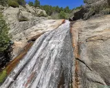 El agua desciende por la roca en la cascada del Hornillo, rodeada de vegetación y rocas graníticas bajo un cielo azul.