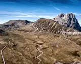 Vasto panorama del **campo imperatore gran sasso** con strade tortuose che si snodano tra montagne brulle e un cielo azzurro intenso.