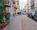 Wjazd do Wiednia samochodem osobowym. Ulica z kawiarniami, sklepami i zielenią, z zaparkowanymi autami i przechodniami.