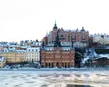 Vinter i Stockholm med snöklädda tak och isig vattenyta. Vackra byggnader reser sig mot en klar himmel.