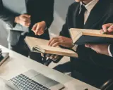 Trzy osoby w garniturach analizują dokumenty i książki przy biurku z laptopem i wagą sprawiedliwości.