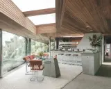 Interno della Sheats Goldstein Residence con soffitto in legno, ampie vetrate, zona pranzo con tavolo in vetro e sedie in pelle arancione, e un bancone bar in cemento con sgabelli.