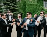 Groomsmen in black suits and cream ties, with the groom in a navy suit, all sporting sunglasses and a playful mood. Do groomsmen match the groom? Not exactly, but they're all dressed to impress!