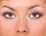 Primo piano di un viso femminile con occhi piccoli e verdi, ciglia lunghe e trucco viola sulle palpebre.