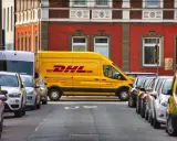 Żółta furgonetka DHL stoi zaparkowana na ulicy, obok innych samochodów. To miejsce, do którego pracują kurierzy DHL, by dostarczać paczki.