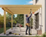 Pose d'une fixation de tonnelle sur terrasse. Un ouvrier ajuste la structure en bois avec des outils.