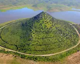 El **cerro Masatrigo**, cubierto de vegetación, se alza majestuoso junto a un embalse. Una carretera serpentea a su alrededor, conectando con un puente.