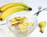 Kawałki skórek bananów w wodzie, przygotowywane jako nawóz z banana. Obok leżą banany i nożyczki.