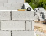 Miarka budowlana na murze z bloczków betonowych. W tle stosy materiału budowlanego. Ile bloczków betonowych na m2?