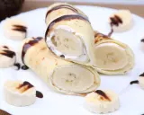 Rolowane naleśniki z bananem i kremem, polane czekoladą. Na talerzu ułożone są też kawałki banana z polewą.
