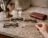 Kobieta z twarzą diamentową wybiera okulary. Na stole leżą różne modele, portfel i biżuteria.