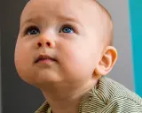 Occhi azzurri di un neonato di 10 mesi che guarda in alto, con un'espressione curiosa. Indossa una maglietta a righe gialle e nere.