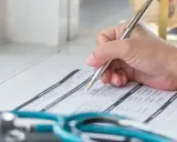 Dłoń pisząca długopisem na formularzu medycznym, obok stetoskopu.