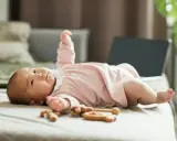 Urocze niemowlę patrzy do góry, leżąc na łóżku w różowej bluzeczce. Obok leżą drewniane zabawki.