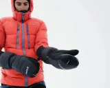 Mężczyzna w pomarańczowej kurtce puchowej zakłada rękawicę z jednym palcem inaczej, przygotowując się na zimową wyprawę.