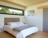 Chambre couleur taupe avec un grand lit blanc, une tête de lit capitonnée et une vue sur la nature par la fenêtre.