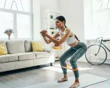Femme faisant des squats avec une bande de résistance pour affiner ses jambes, dans un salon lumineux.