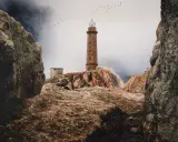 Leuchtturm auf felsiger Küste der Costa da Morte, Vögel ziehen über neblige Berge.
